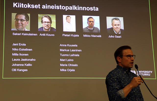 Henkilö puhuu mikrofoniin, taustalla heijastettuna kuvia ja nimiä.