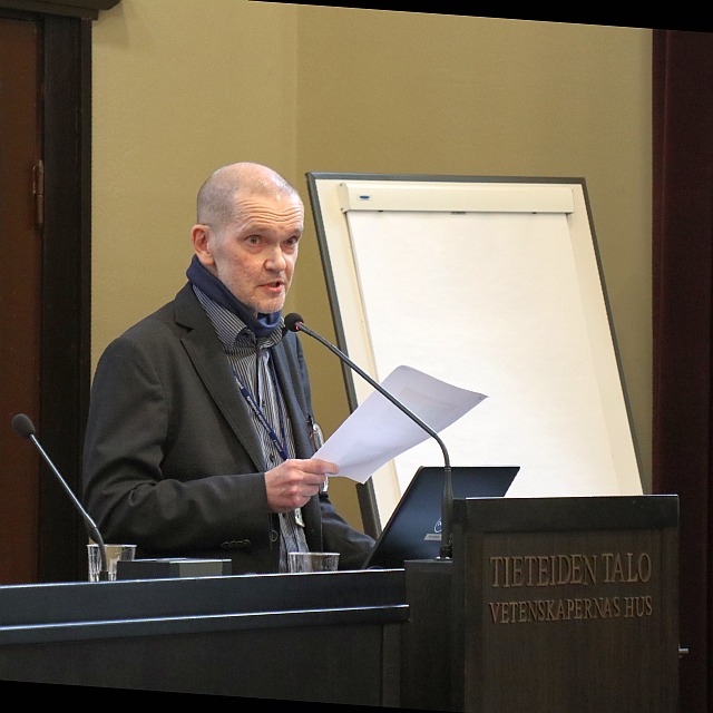 Kokoustila, puhujakorokkeen takana mies, jolla päällään puku ja joka katsoo yleisöön kädessään paperiarkki.
