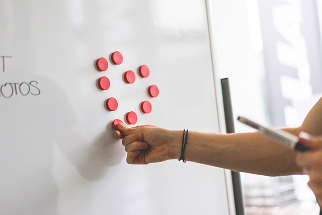 A whiteboard with ten pink magnets arranged in the shape of a heart. A hand places the final magnet in position.