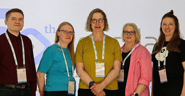 Viisi tapahtuman osallistujaa poseeraa yhteiskuvassa konferenssilavan edessä. Kaikilla on tapahtuman nimikortit kaulassa.
