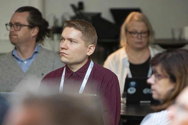 Seminaariyleisöä kuuntelemassa esitystä konferenssi- tai seminaaritilaisuudessa. Kuvan keskellä istuu osallistuja kaulassaan tapahtuman nimikorttinauha.