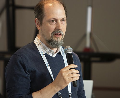 Konferenssiosallistuja pitää kädessään mikrofonia ja puhuu yleisölle. Kaulassa on tapahtuman nimikorttinauha, ja taustalla näkyy sisätilan kokousympäristöä.