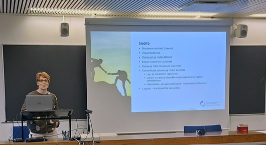 Luentosali, jossa silmälasipäinen puhuja vasemmalla tietokoneen ääressä. Ruudulle on heijastettu esitysdia.