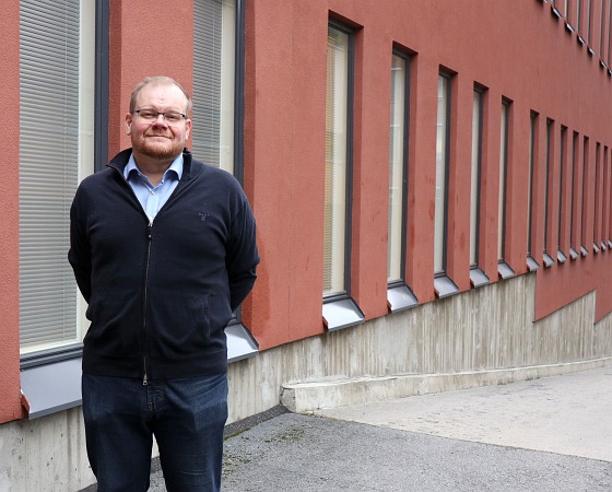Etualalla Matti Heinonen seisoo ulkona, taustalla ikkunaseinä ja ajoluiska